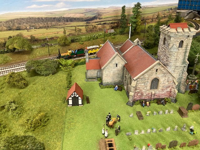 A model railway of Hampton Lovett Church, surrounded by lush green fields.